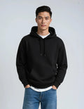 A man wearing a black pullover hoodie with a front kangaroo pocket and drawstrings, layered over a white t-shirt and paired with blue jeans, standing against a light gray studio background.