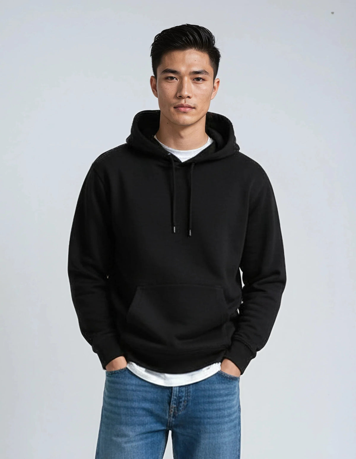 A man wearing a black pullover hoodie with a front kangaroo pocket and drawstrings, layered over a white t-shirt and paired with blue jeans, standing against a light gray studio background.